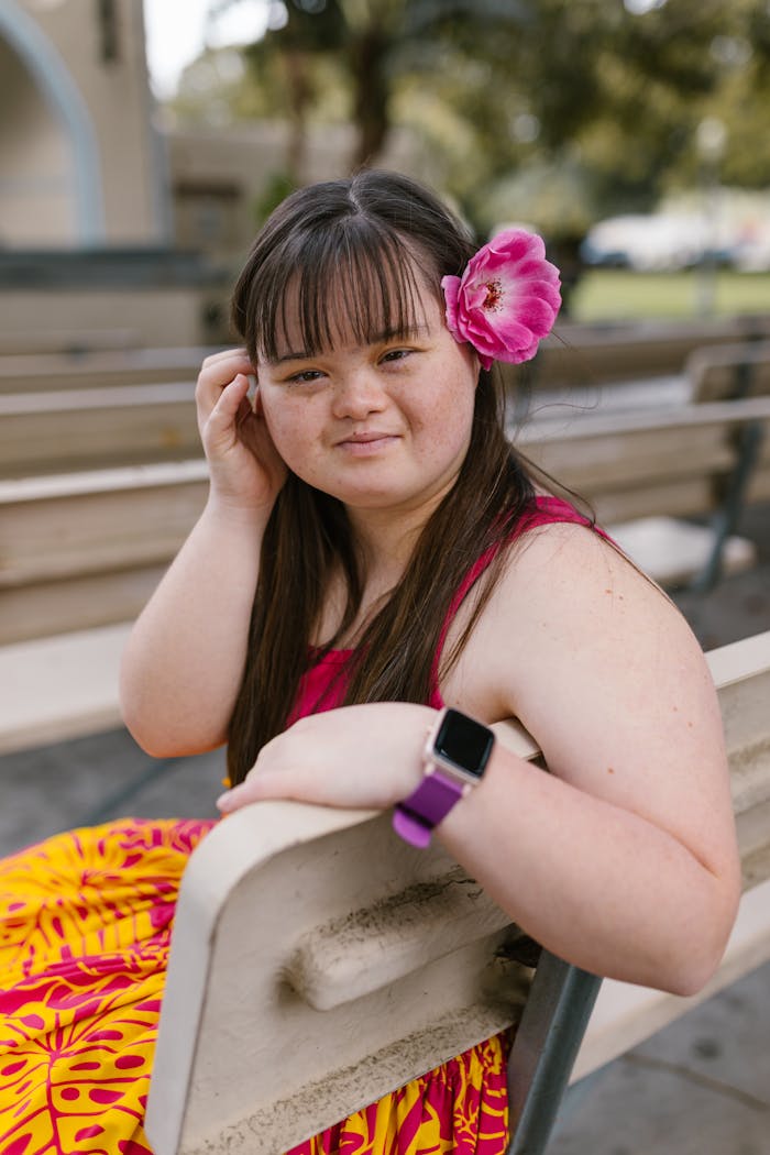 digital-01 Young woman with Down syndrome wearing floral dress, sitting on bench, smiling with flower in hair.